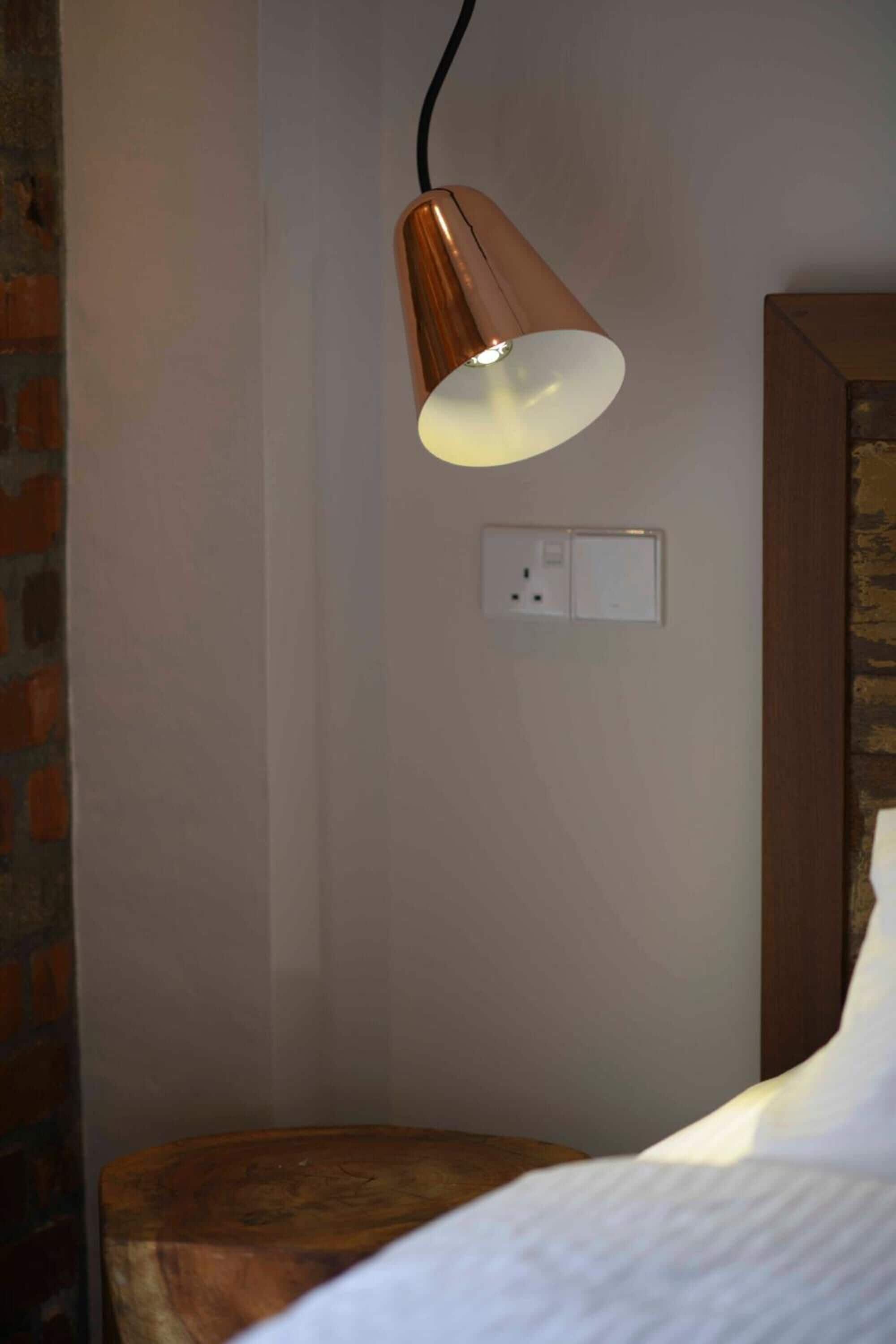 A single bed with a wooden headboard, a wall-mounted lamp, and a power outlet on a plain wall.