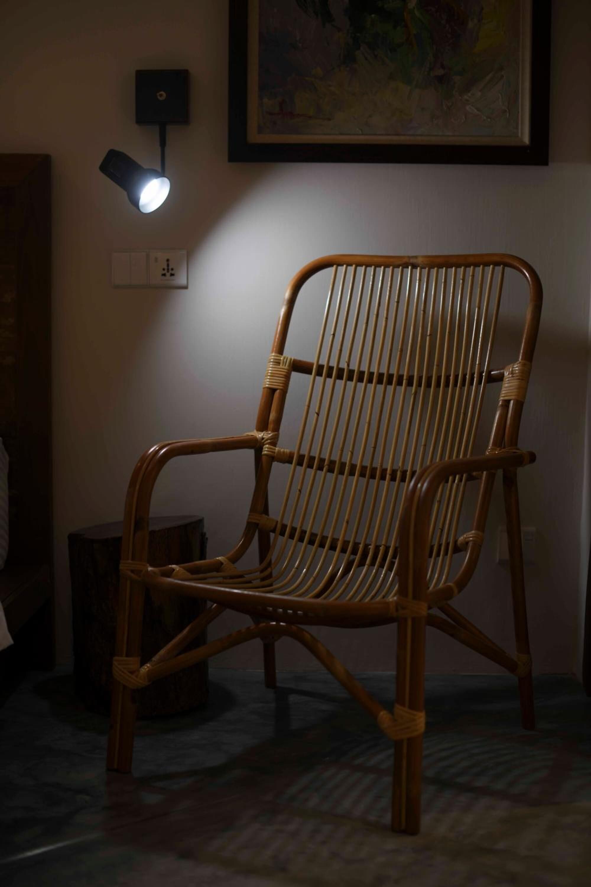 A wicker chair with a backrest, positioned in a room with a wall-mounted light fixture and a framed artwork above it.