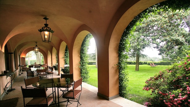 Un patio couvert, aménagé avec des tables et des chaises, des arcades et offrant une vue sur une pelouse et un jardin bien entretenus.