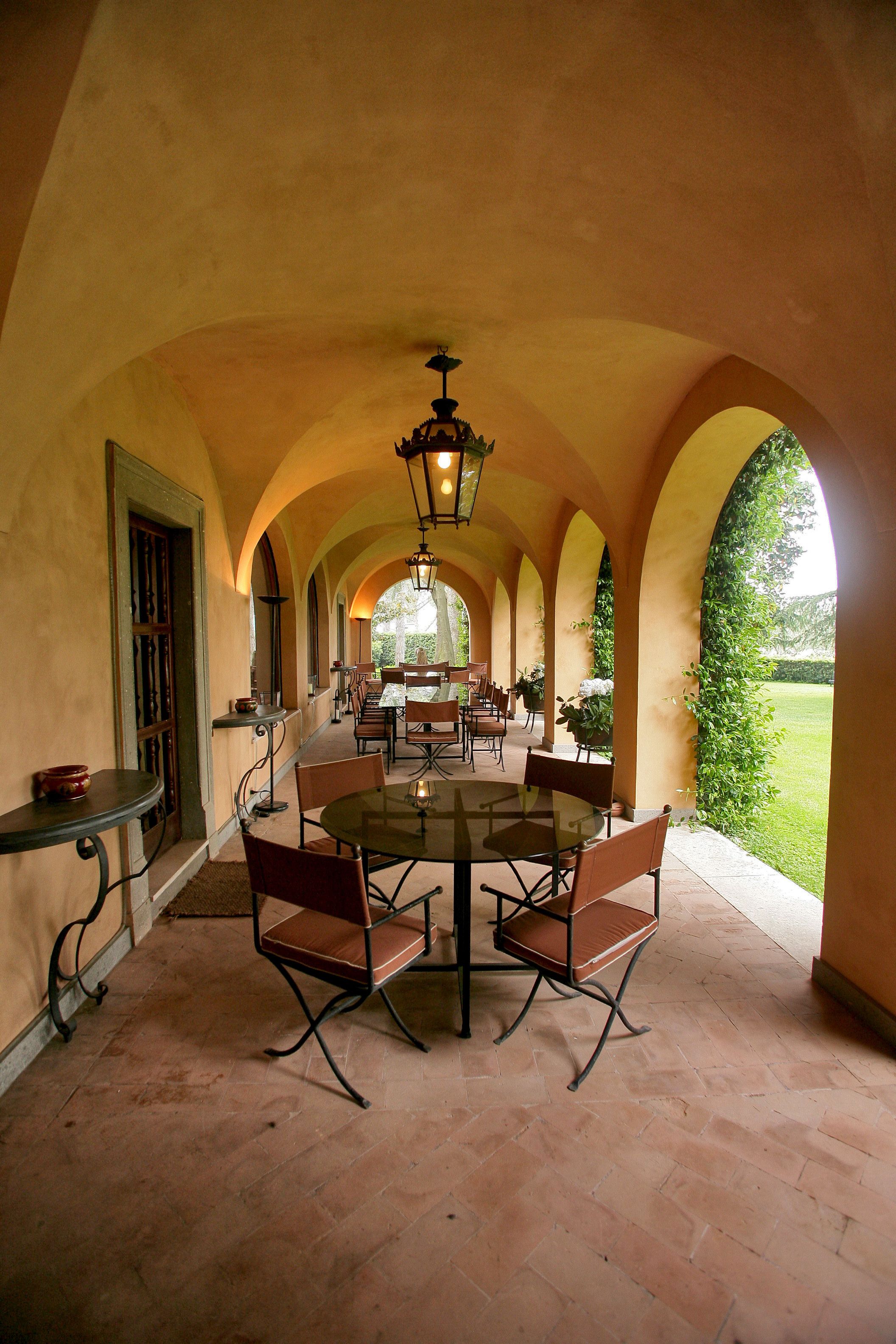 Une terrasse extérieure avec une longue table et des chaises, entourée d’arcades et offrant une vue sur la verdure.