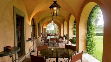 Une terrasse extérieure avec une longue table et des chaises, entourée d’arcades et offrant une vue sur la verdure.