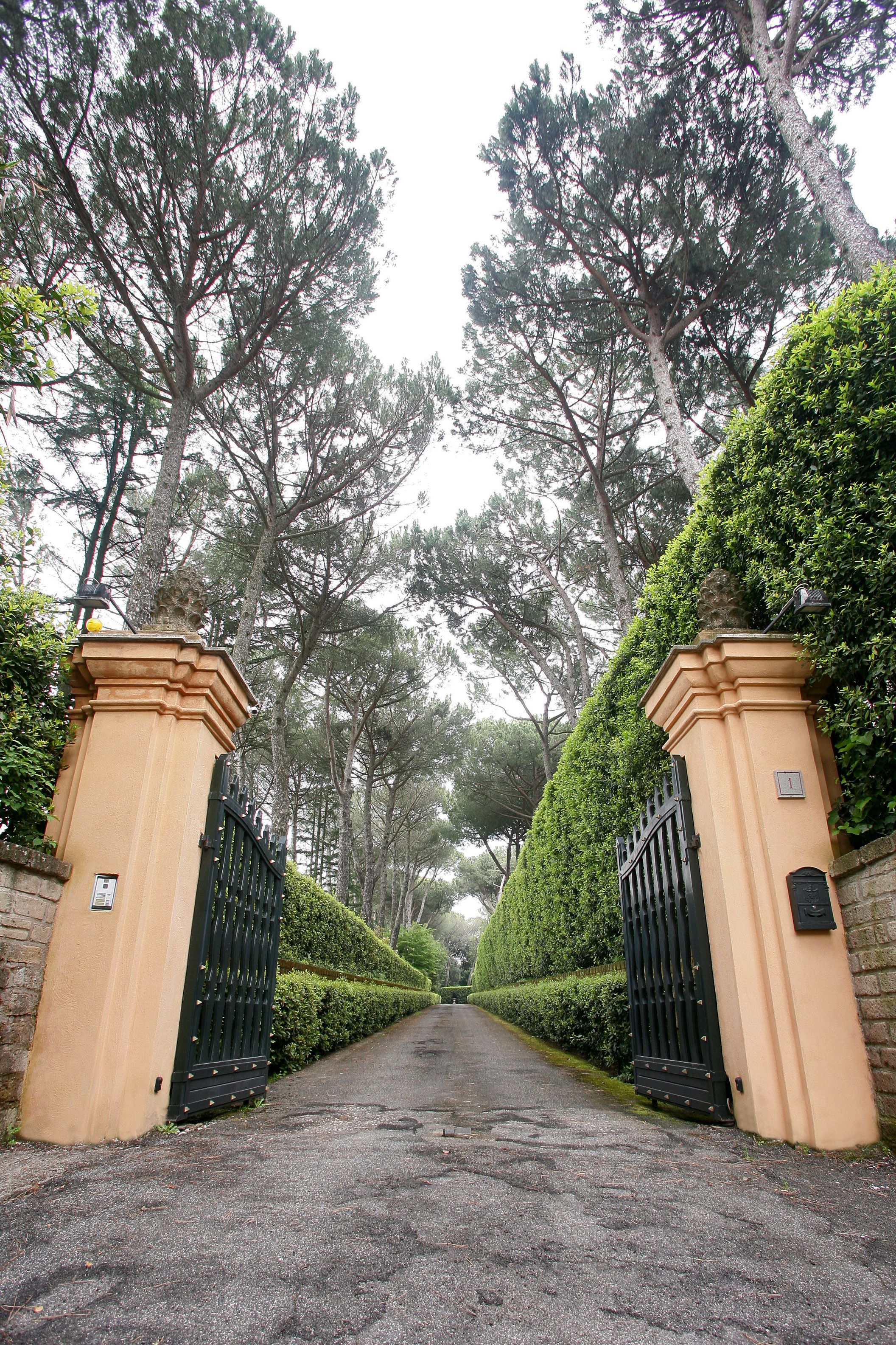 A gated entrance with a paved path leading through a landscaped area with tall trees and trimmed hedges.