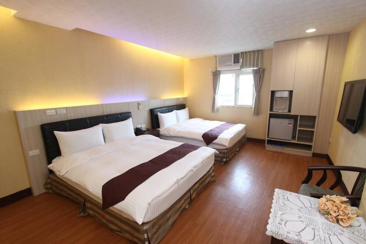 A hotel room with two beds, a TV, a small kitchenette, and a window with curtains.
