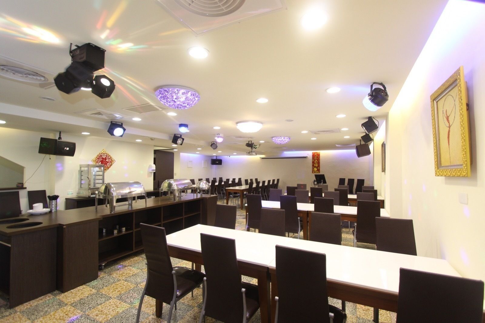 Un salón de conferencias con varias mesas y sillas, equipado con luces en el techo y un proyector.