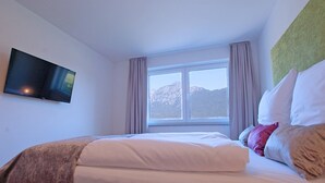 Hotellihuone, jossa on sänky, seinälle asennettu televisio, ikkuna verhoilla ja näkymä vuorille.