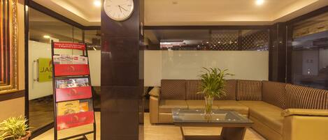 A lobby with a clock, a sofa, a coffee table, a magazine rack, and a potted plant.