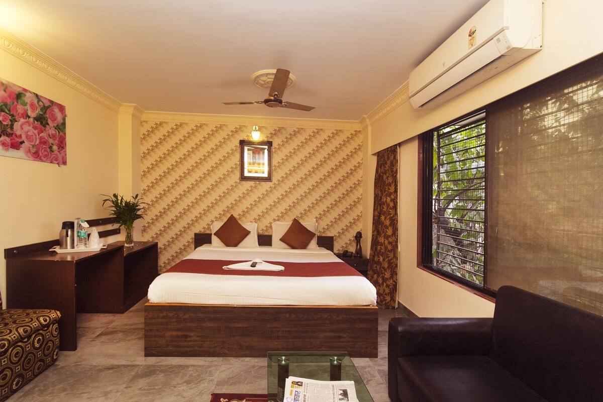 Habitación de hotel con cama, escritorio, silla, mesa de vidrio y ventana con vista a árboles.