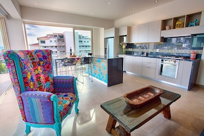 A modern kitchen with a large island, a dining area with colorful chairs, and a view of the city through large windows.