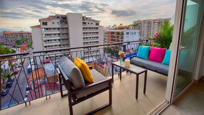 Un balcón con sofá, una silla, una mesa y vistas a un paisaje urbano con edificios y una carretera.