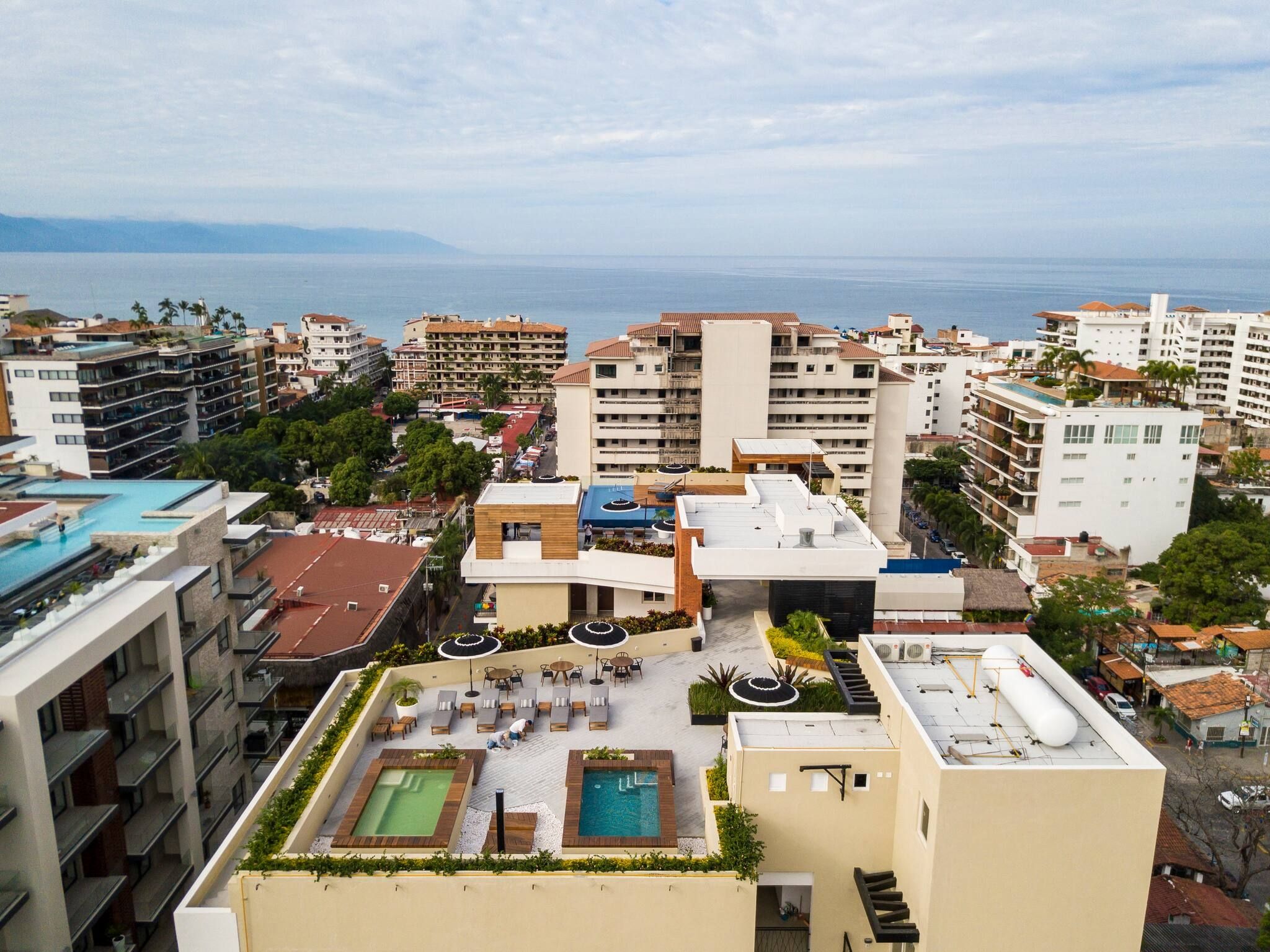A rooftop with a swimming pool and outdoor seating, surrounded by multi-story buildings and a view of the sea.