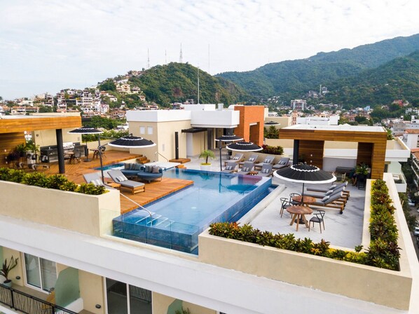A rooftop pool area with lounge chairs and umbrellas, surrounded by buildings and a mountainous backdrop.