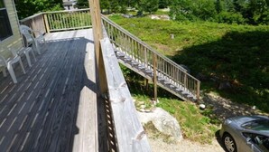 Una terraza de madera con mobiliario exterior, una escalera que lleva a una zona cubierta de césped y un coche aparcado cerca.