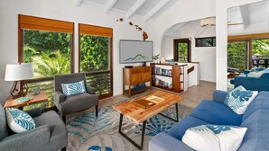A spacious living room with a large window, a blue sofa, a grey armchair, a wooden coffee table, and a bookshelf. - Kahala at Poipu Kai #121 By Parrish Kauai - walk to Poipu Beach & great for hone (Koloa)