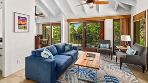 A spacious living area with a blue sofa, a wooden coffee table, and a grey armchair. There is a ceiling fan, a large window with a view of greenery, and a poster on the wall. - Kahala at Poipu Kai #121 By Parrish Kauai - walk to Poipu Beach & great for hone (Koloa)