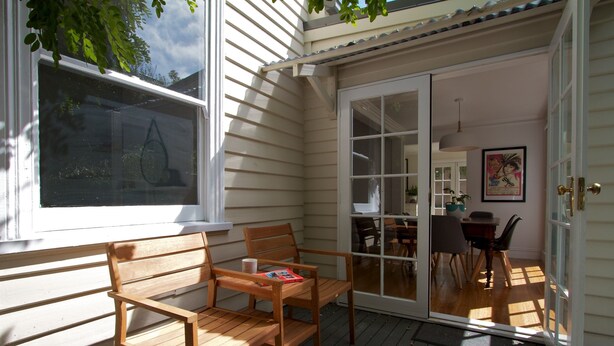 Una terraza de madera con mesa y sillas, una puerta corrediza de vidrio que da a un comedor interior y una ventana con persianas.