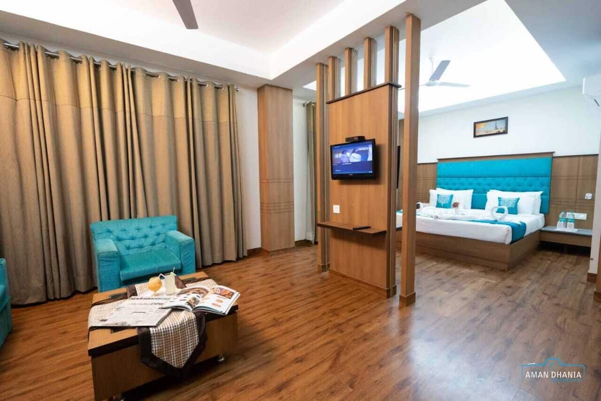 A hotel room with a wooden floor, a teal sofa, a bed with a blue headboard, a TV on a wooden stand, and a window with curtains.