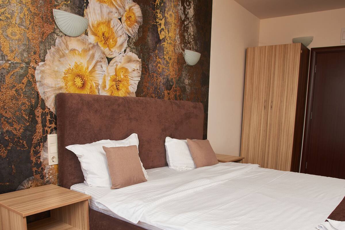 A bedroom with a large bed, a wooden nightstand, a wall-mounted lamp, and a wardrobe.