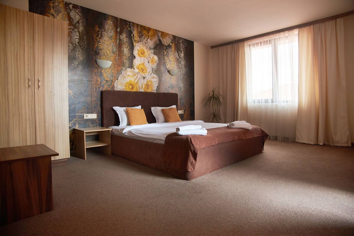 A bedroom with a large bed, a wooden wardrobe, a small table, and a wall with a floral mural.
