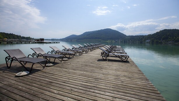 A wooden pier with rows of empty lounge chairs facing a calm lake, surrounded by hills. - See & GenussHotel Silvia (Sankt Kanzian am Klopeiner See)