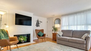 Un salon doté d’une cheminée, d’un téléviseur à écran plat fixé sur un mur en briques blanches, d’un fauteuil en cuir marron, d’un canapé gris et d’une grande fenêtre ornée de rideaux légers.