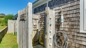Un espace de douche extérieur en bois, équipé d’un tuyau d’arrosage et d’une bouteille suspendue au mur.