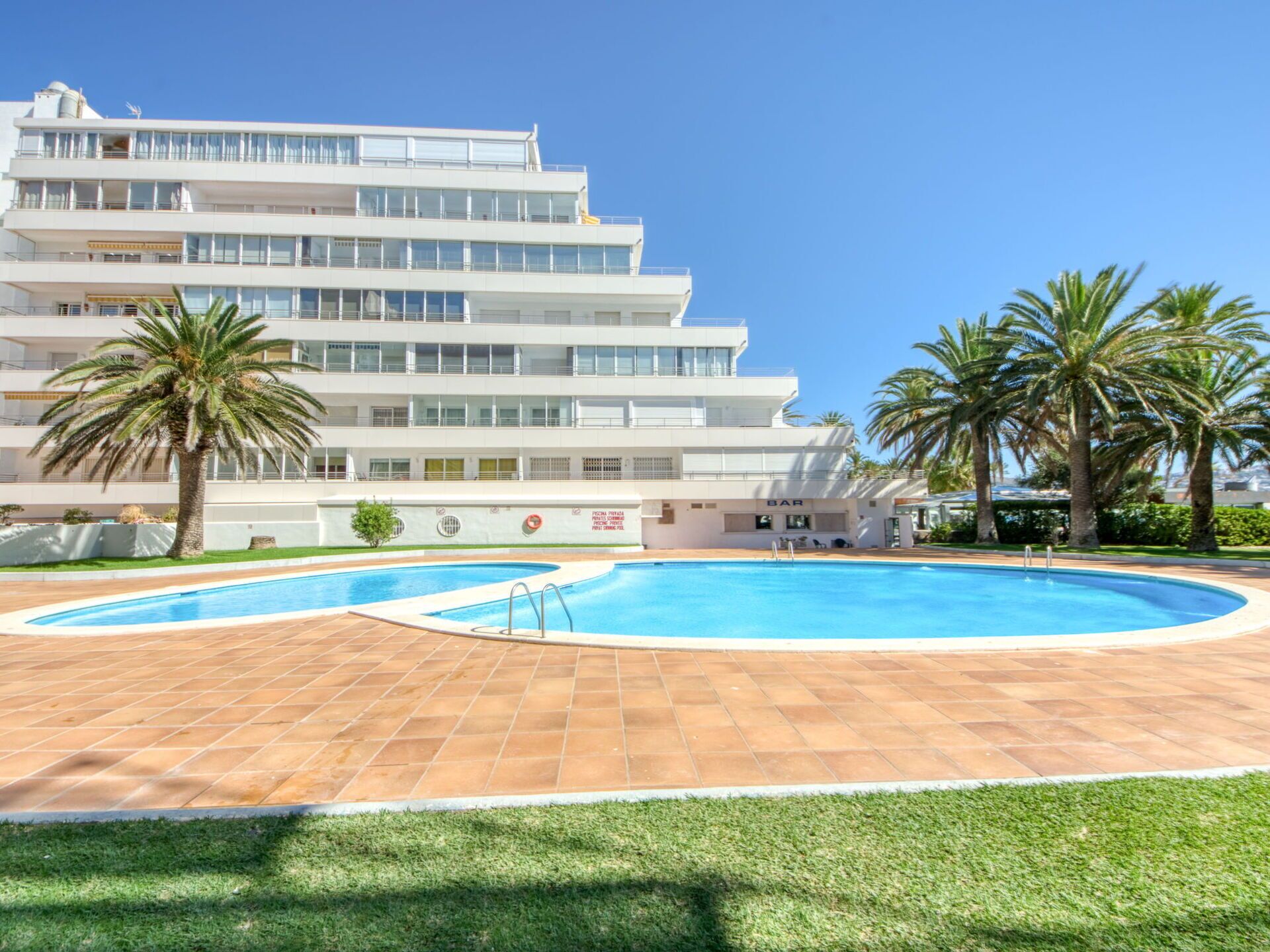 A modern hotel with a pool area, palm trees, and a clear blue sky.