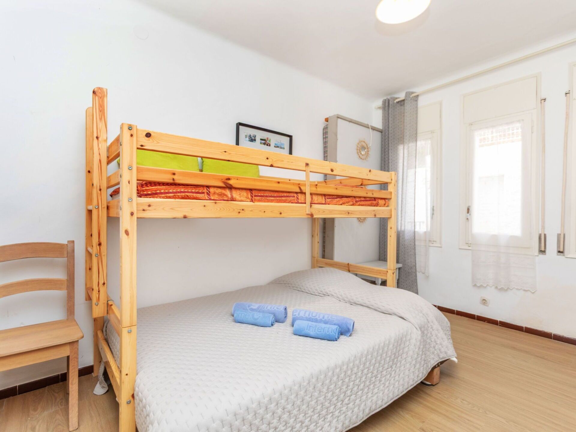 Ein Stockbett mit Holzrahmen, darunter ein Einzelbett, ein Stuhl, ein kleiner Tisch und ein Fenster mit Vorhängen.