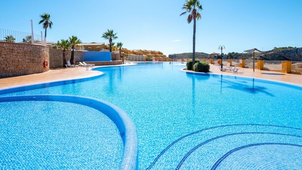 A large, clear blue swimming pool with a curved edge, surrounded by a stone wall and palm trees. - Beautiful apartment for 4 people with WIFI, A/C, pool, TV and patio (Teulada)