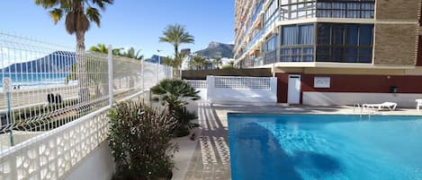 A hotel pool area with a clear view of the ocean, a palm tree, and a building with balconies.