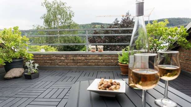 Une terrasse sur le toit, avec un mur en pierre, des plantes en pot, une assiette de fruits secs et deux verres de vin blanc.