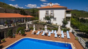 Un espace aménagé au bord de la piscine, avec des chaises longues et des parasols, un mur en pierre et un bâtiment doté d’un toit en tuiles.
