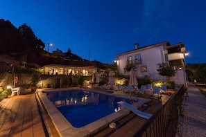Piscina con sillones de descanso y sombrillas, rodeada por un muro de piedra y un edificio con iluminación exterior.