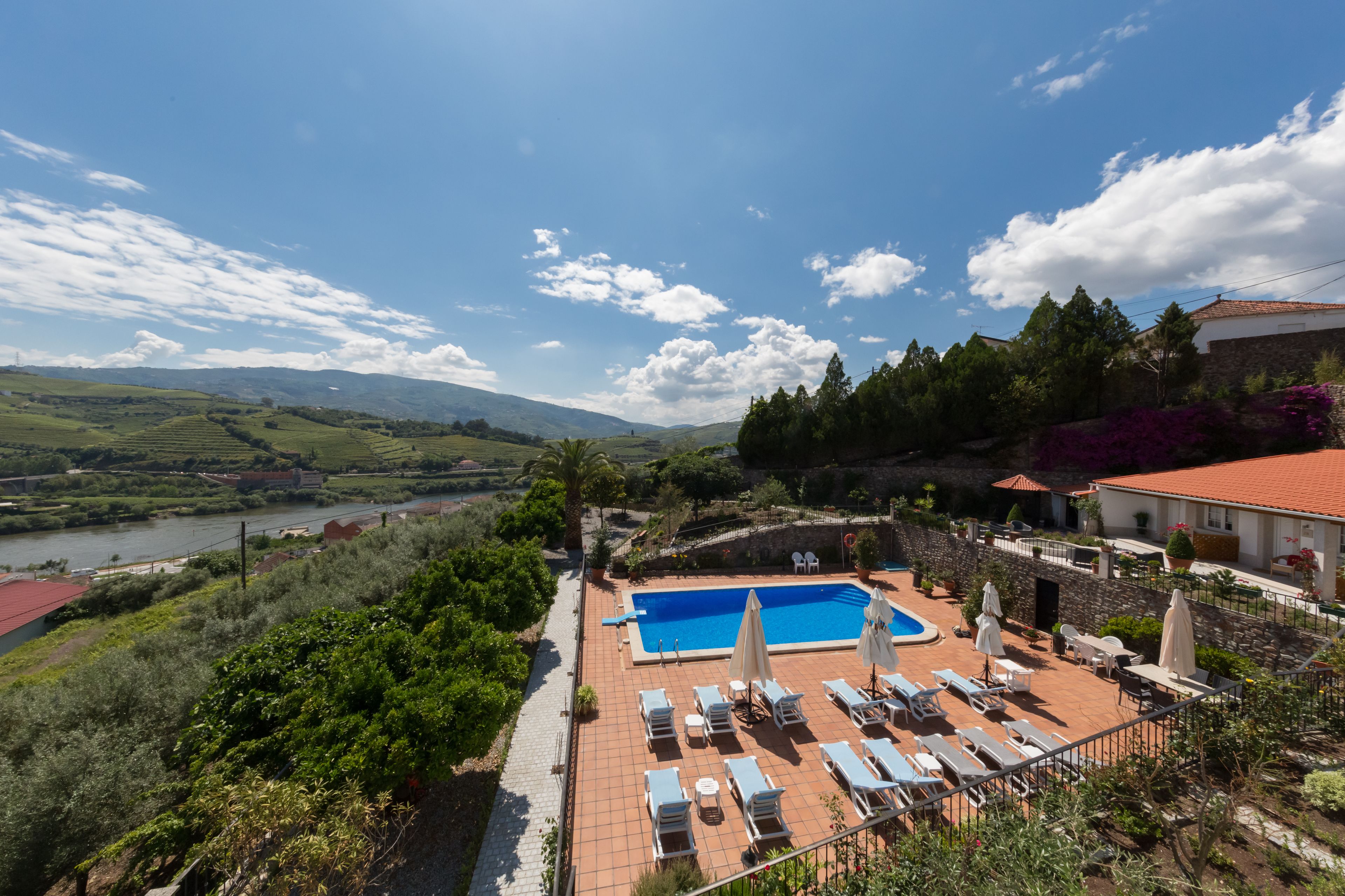 A pool area with lounge chairs and umbrellas, surrounded by greenery and a view of a river and hills.