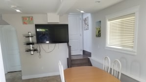 Un comedor con mesa de madera y sillas blancas, un televisor montado en la pared y una ventana con persianas.