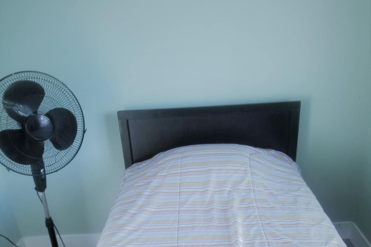 Un lit simple avec une couvre-lit à rayures, un ventilateur sur pied et une serviette blanche posée sur le lit.