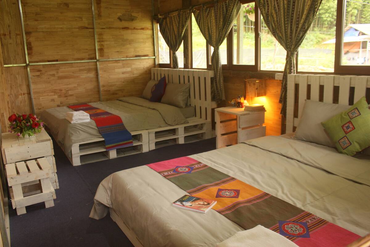 A rustic wooden room with two beds, each with a unique patterned bedspread. There are wooden nightstands with lamps, and a small table with a vase of flowers.