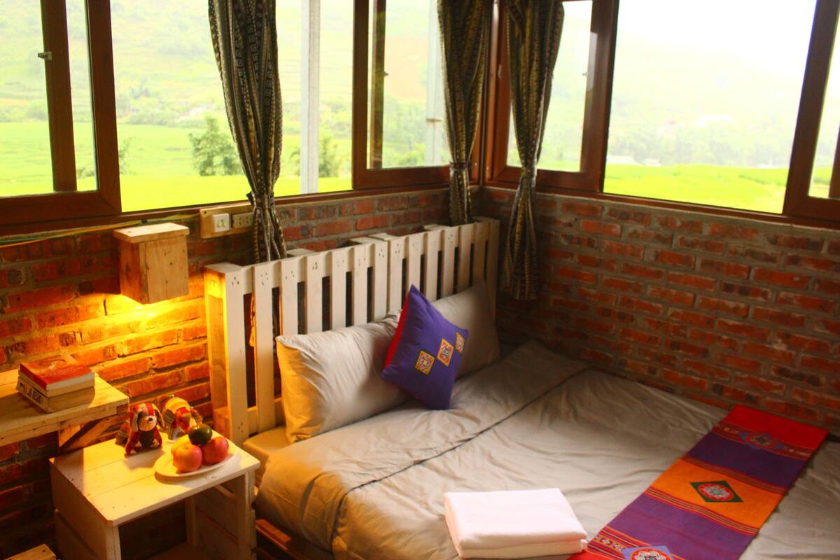 A cozy room with a brick wall, a bed with a colorful blanket, a small table with a teddy bear and apples, and a window with curtains.