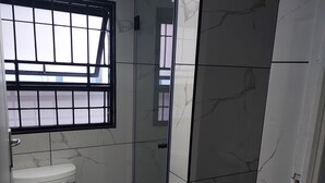 A bathroom with a toilet, a glass-enclosed shower, and a window with bars. - Freedom Media Guest House (Gqeberha (Port Elizabeth))