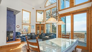Amplia sala de estar con mesa de comedor de vidrio, sillas de madera, chimenea y grandes ventanales con vistas al exterior.