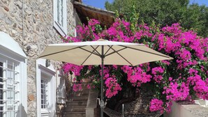 A stone building with a white door and window, a table with a chair under a beige umbrella, and pink bougainvillea flowers. - Alex's traditional summerhouse (Hydra)