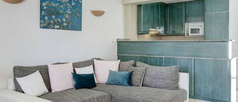 A modern living room with a grey and white sofa, a woven basket, and a kitchen area with teal cabinets and a microwave.