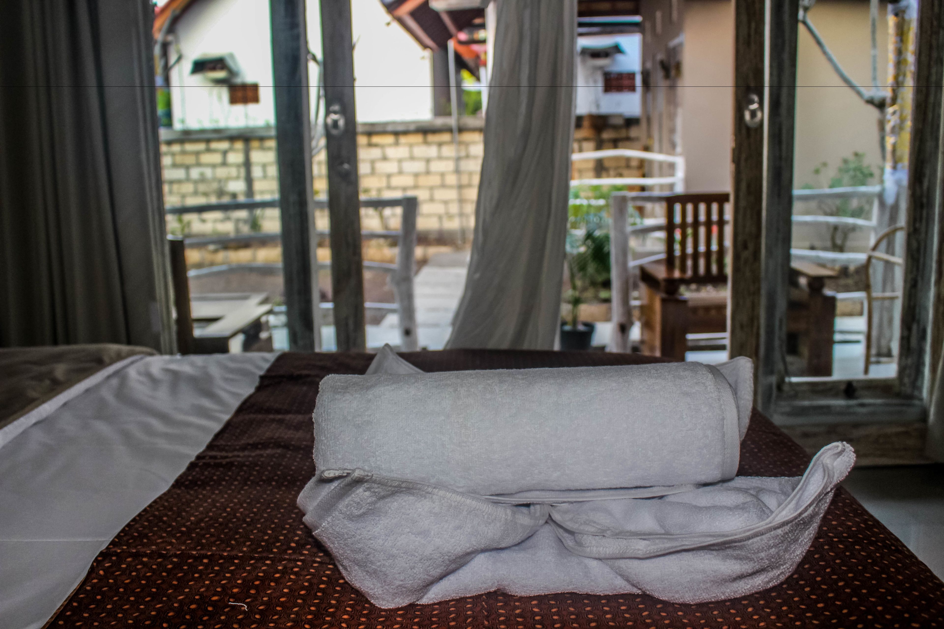 A bed with a folded towel and a polka dot blanket, a wooden bench, and a view of a building through a window.