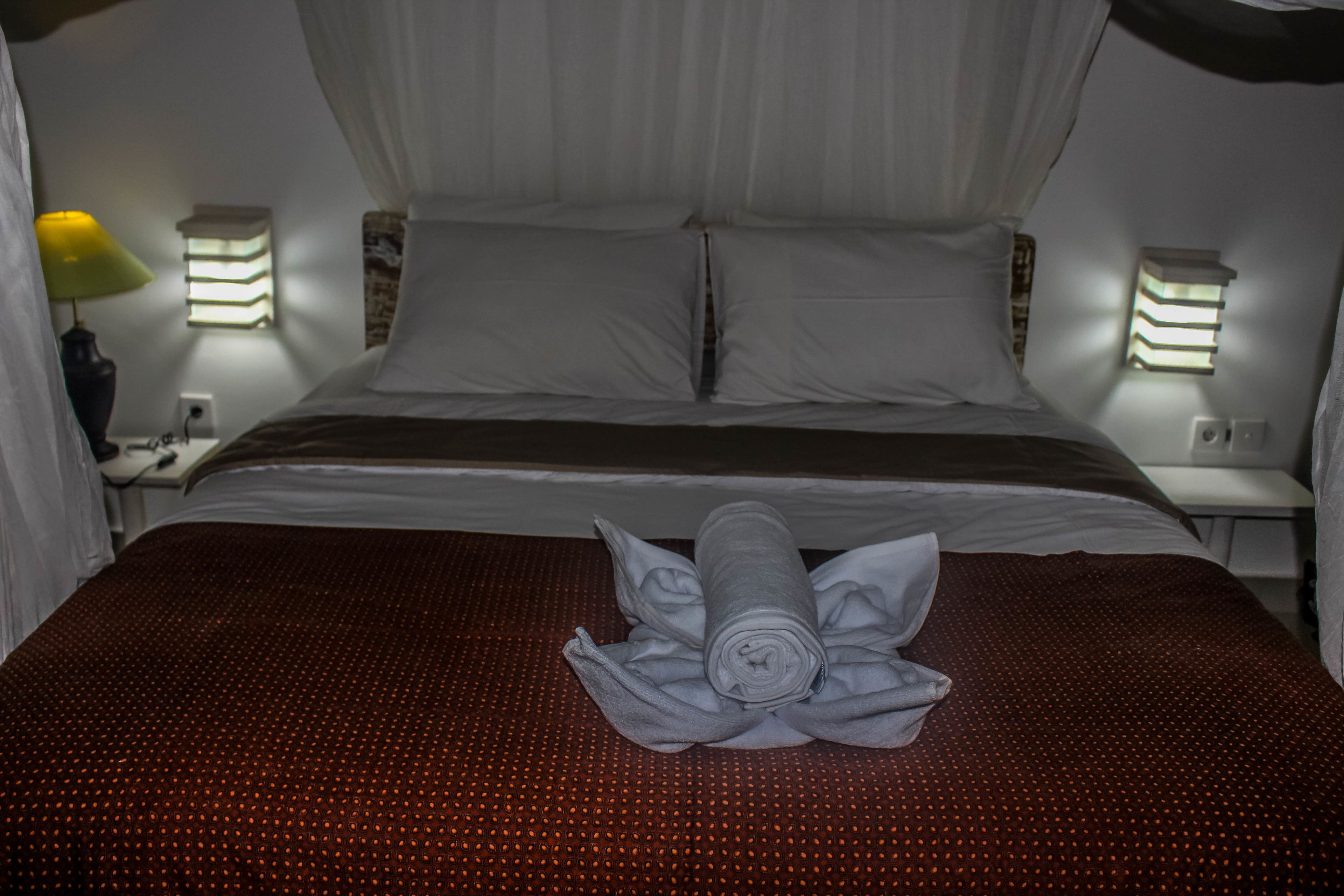 Een net opgemaakt bed met een opgerolde handdoek in het midden, twee bedlampen en een wandlamp.