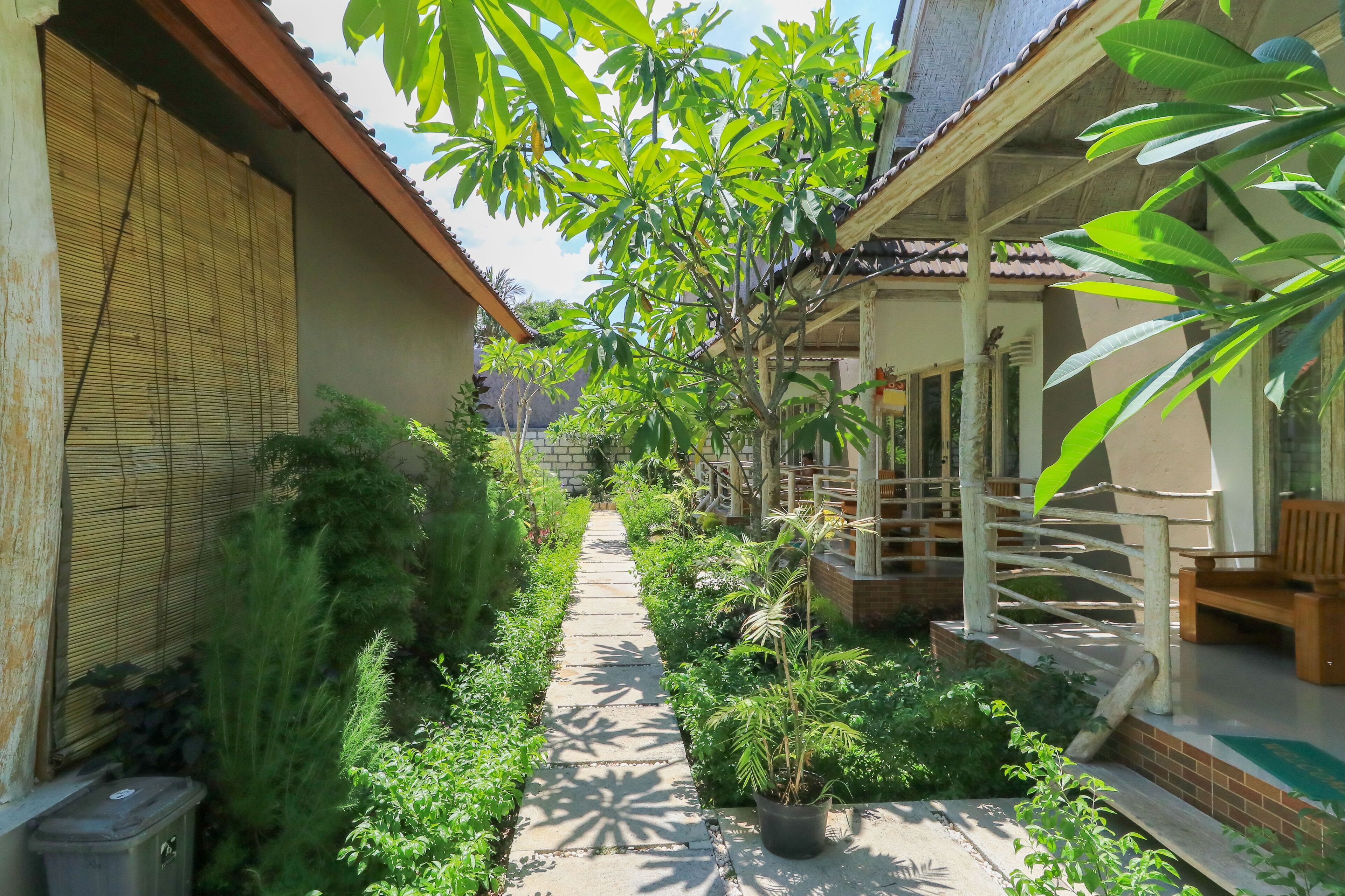 A narrow pathway flanked by lush greenery and traditional architecture, leading to a wooden porch with a bench.