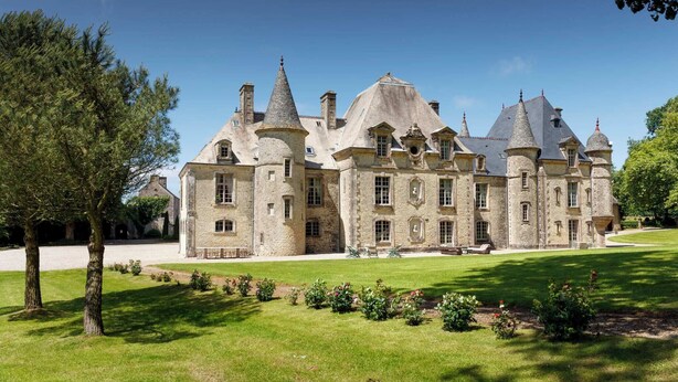 A large, historic stone building with multiple towers and turrets, surrounded by a well-maintained lawn and trees.