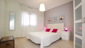 Un dormitorio con una cama grande, cortinas blancas, una mesita de noche con lámpara y una pared decorada con marcos.