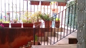 Un espacio de estar al aire libre con techo, con una mesa de madera y bancos, rodeado de plantas en macetas y una barandilla de metal.