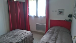 A bedroom with two beds, red curtains, a window with sheer curtains, a wall-mounted heater, and a small table with a lamp. - RENTAL DOMAINE DE LA BAUME - "LE MOULIN" (Cendras)