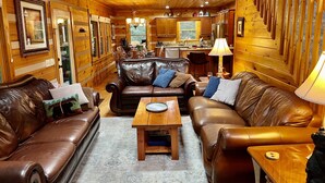 A cozy log cabin interior with leather sofas, a wooden coffee table, and a kitchen area visible through an open door. - Luxury Cabin with Mountain Views, Fire pit, Hot tub and more ! (Boone)