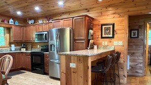 A wooden cabin kitchen with a stainless steel refrigerator, microwave, and stove. There are wooden cabinets, a granite countertop, and bar stools. - Luxury Cabin with Mountain Views, Fire pit, Hot tub and more ! (Boone)